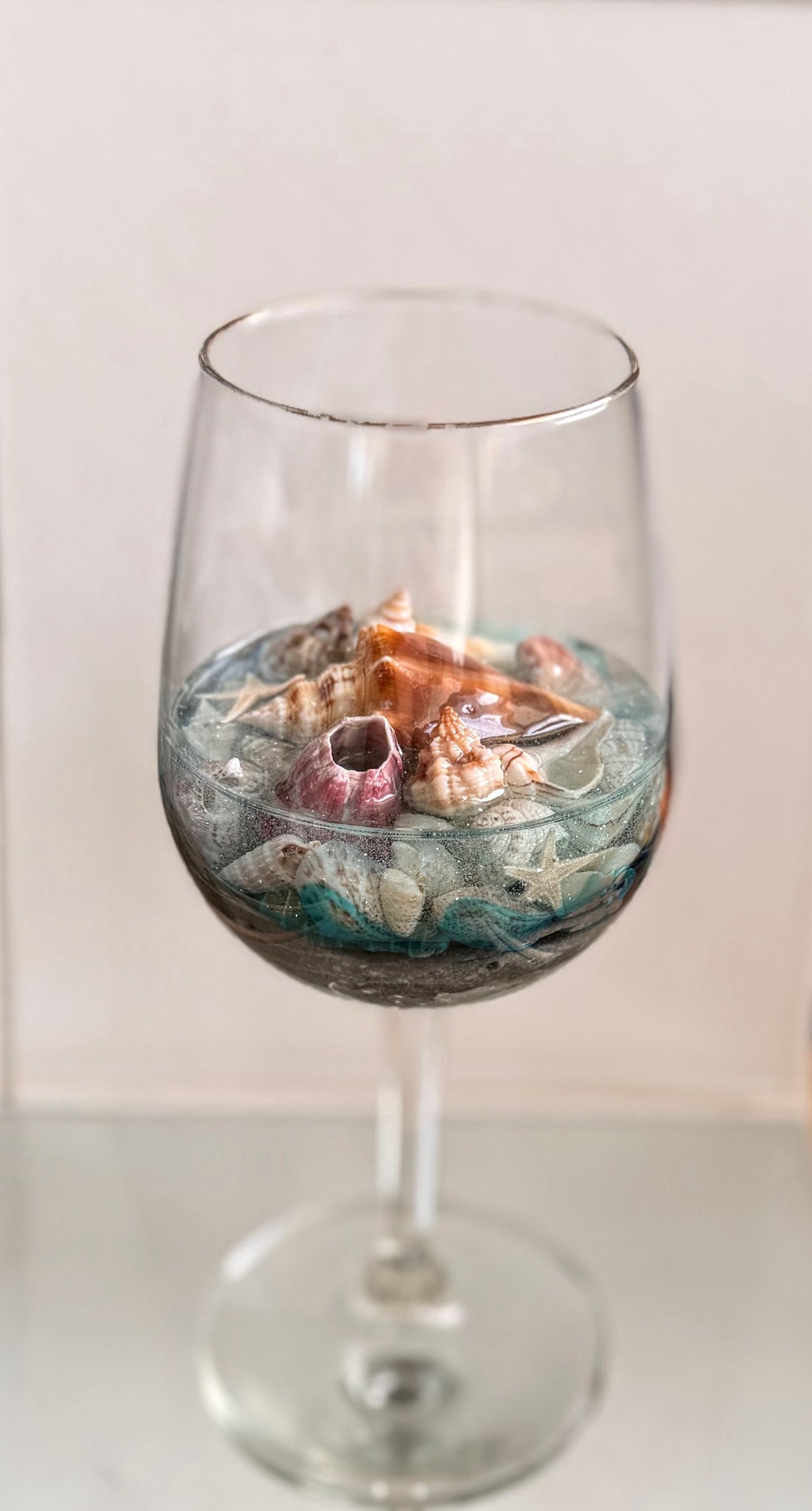 A clear wine glass filled with blue-green water and natural seashells, photographed against a plain light background.